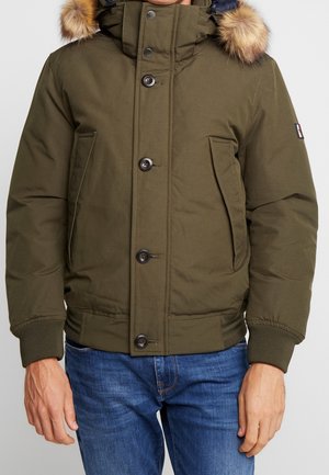 Homme portant une veste matelassée vert olive avec capuche bordée de fourrure, devant avec boutons et fermeture éclair, et un jean bleu.