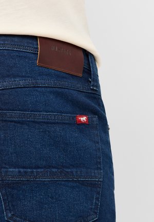Dark blue denim jeans with a brown leather label embossed with "MUSTANG" and a small red tag featuring a horse logo on the back pocket.