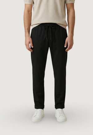 Uomo che indossa pantaloni neri con coulisse risvoltati alle caviglie, scarpe da ginnastica bianche e una camicia beige a maniche corte, in piedi davanti a uno sfondo semplice.