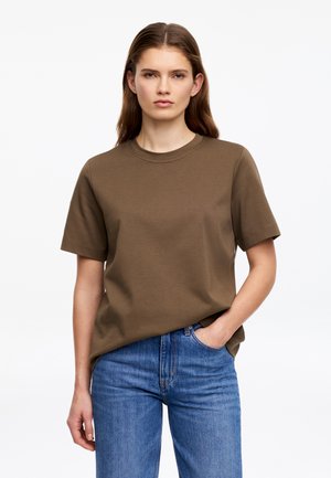 Kobieta o brązowych włosach nosi prostą oliwkowozieloną koszulkę z krótkim rękawem i niebieskie dżinsy, stoi na tle jednolitego białego tła.