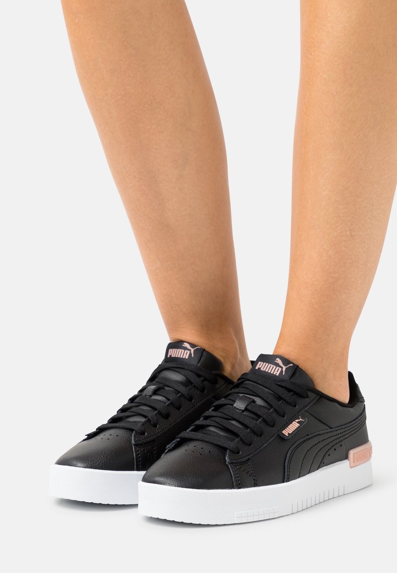 Baskets en cuir noir avec semelle en caoutchouc blanche, présentant une finition texturée, un design à lacets et un accent rose sur le côté.