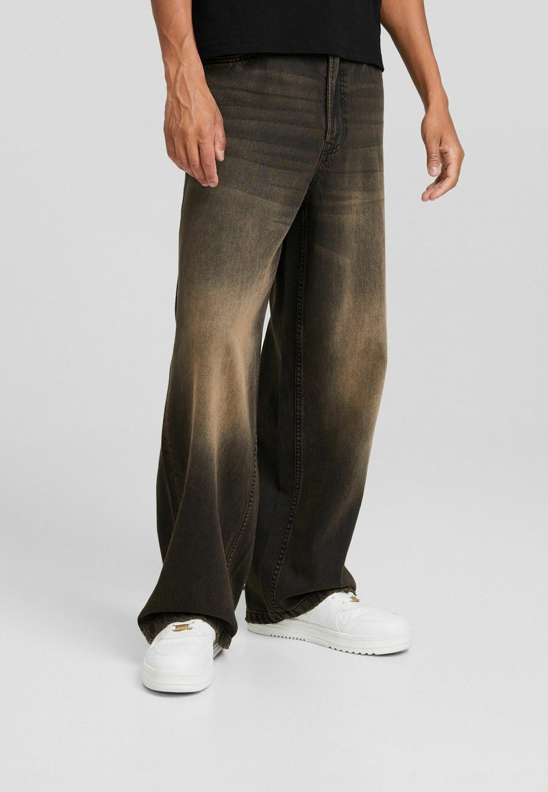 Personne portant un jean noir délavé à jambes larges et des baskets blanches, debout devant un fond gris clair uni.
