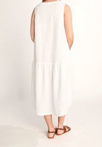 Weißes, ärmelloses Leinenkleid mit Stufendesign, das knapp unter die Knie fällt. Dazu braune Sandalen, die eine entspannte Silhouette zeigen.