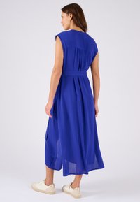 Abito maxi di un blu vibrante, realizzato in tessuto fluente. Presenta maniche a sbuffo, una vita cintrata con una cintura e pieghe sul retro. Abbinato a sneakers leggere.