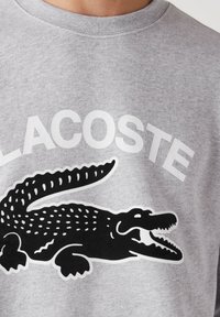 Gros plan sur un sweat-shirt gris avec un logo crocodile noir et le mot « LACOSTE » en grandes lettres blanches au-dessus.