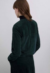 Chaqueta de pana verde oscuro con una superficie texturizada, que presenta un cuello alto negro y un diseño recortado con un ajuste relajado.