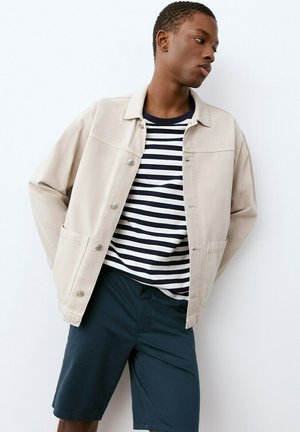 Giovane uomo che indossa una giacca beige sopra una maglietta a righe bianco-nera e pantaloncini blu scuro, in piedi contro uno sfondo bianco.