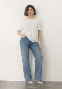 T-shirt bianca con decorazioni, abbinata a jeans a gamba larga blu chiaro e tacchi a punta color nude, su uno sfondo semplice.