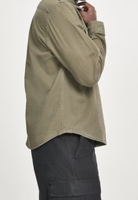 Olivgrön långärmad skjorta med knäppt krage och rak skärning, tillsammans med svarta cargobyxor med sidofickor.