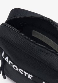 Zaino in tessuto nero con chiusura a zip, caratterizzato da un prominente logo bianco "LACOSTE" e tessuto testurizzato. All'interno è presente una piccola tasca.