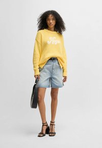 Maglione giallo a maglia con logo "Hugo", abbinato a pantaloni corti in denim azzurro chiaro. Il modello indossa tacchi neri con cinturini e tiene una borsa nera.