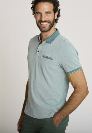 Homme souriant aux cheveux foncés bouclés portant un polo gris clair avec une bordure bleu sarcelle et un pantalon vert, debout avec une main dans la poche.