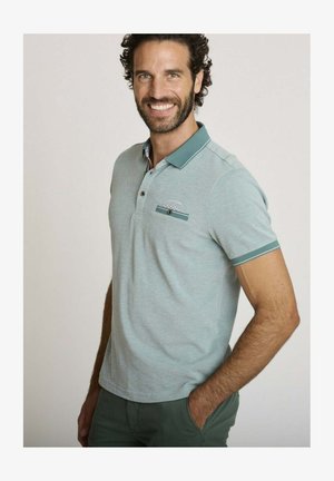 Homme souriant aux cheveux foncés bouclés portant un polo gris clair avec une bordure bleu sarcelle et un pantalon vert, debout avec une main dans la poche.