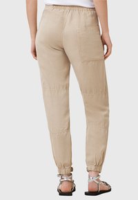 Béžové jogger kalhoty s elastickým pasem, bočními kapsami, zúženými manžetami a prošívanými detaily. V kombinaci se stříbrnými sandály na řemínky.