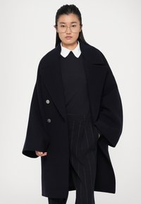 Námořnický vlněný kabát s širokým límcem, dvojitými knoflíky a oversized designem, kombinovaný s černým svetrem a kalhotami s proužky. Průhledné brýle.