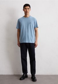 Lichtblauw katoenen t-shirt met een klein logo op de borst, gecombineerd met zwarte broek en zwarte schoenen. Model staat tegen een gestructureerde witte muur.