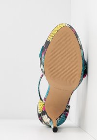 Chaussure à talon haut colorée unique avec motif peau de serpent, bout pointu et semelle texturée, positionnée debout contre un mur blanc.