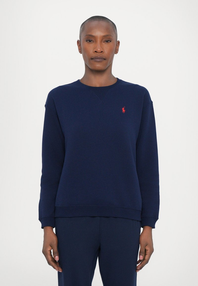 Marineblå crewneck genser laget av mykt stoff, med ribbet halslinning og mansjetter, samt en liten rød logo på venstre bryst.