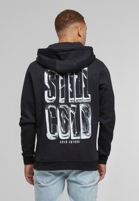 Svart hettegenser med stor hvit trykk på baksiden med teksten "STILL COLD" og "COLD FUTURE." Laget av mykt stoff med avslappet passform.