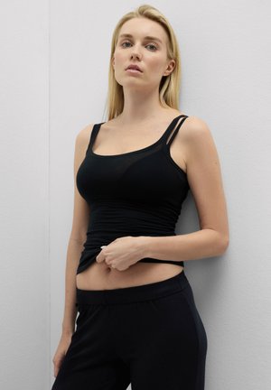 Junge Frau mit blonden Haaren, die ein schwarzes Tanktop und eine Hose trägt, steht vor einer weißen Wand und hebt ihr Oberteil leicht an.