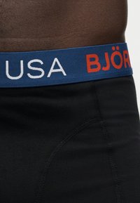 Κοντινό πλάνο ενός ατόμου που φοράει μαύρα εσώρουχα με μπλε ελαστική μέση με λευκό και κόκκινο κείμενο "USA BJÖR".