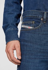 Dark blue denim jeans with yellow stitching, featuring a visible pocket label reading "Diesel Industry." Close-up of a hand on the waistband.