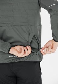 Grå sportsjakke med lynlåslommer i siden og elastiske manchetter, med en let og glat tekstur samt "ENDURANCE" logo ved sømmen.