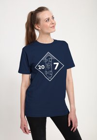 Junge Frau mit langen Haaren in einem Pferdeschwanz, die ein navyblaues T-Shirt mit einem weißen Skateboard-Graphic und den Zahlen 20 und 7 trägt.