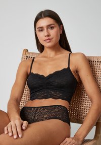 Bralette en dentelle noire avec des fines bretelles et un devant froncé, associé à un slip en dentelle assorti. Les deux présentent des motifs floraux élaborés.