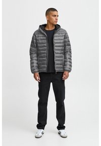 Grå polstret jakke med hette, med et quiltet design. Brukt over en svart skjorte, kombinert med mørke bukser og hvite sneakers.