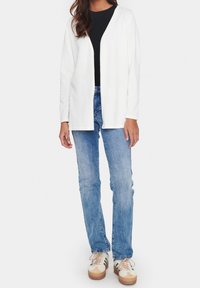 Kvinna som bär en vit öppen kardigan, svart topp, blå jeans och beige sneakers med gröna ränder som står mot en vit bakgrund.