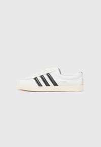Baltas sneakers ar melnām svītrām, noapaļota purngala, teksturēta auduma un bēša gumijas zole. Satur minimālu zīmola nostiprinājumu sānos.
