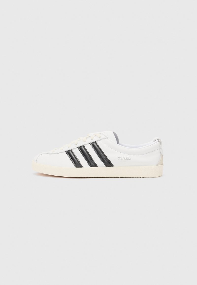 Baltas sneakers ar melnām svītrām, noapaļota purngala, teksturēta auduma un bēša gumijas zole. Satur minimālu zīmola nostiprinājumu sānos.
