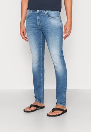 Hellblaue Slim-Fit-Jeans mit einer ausgewaschenen Optik, fünf Taschen und Metallbeschlägen, kombiniert mit schwarzen Flip-Flops auf einem einfarbigen Hintergrund.