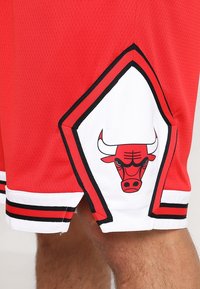 Shorts de basket-ball rouges avec des bordures noires et blanches, arborant un patch avec un logo de taureau sur le côté, au-dessus d'une jambe inférieure visible.