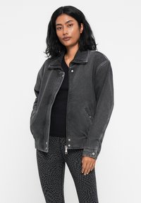 Grå denimjakke med krave, glidelås og sidelommer. Båret over en svart topp og mønstrede leggings. Glatt tekstur og avslappet passform.