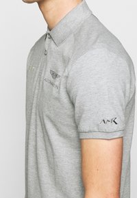 Šedé polo tričko vyrobené z bavlněné směsi, s límečkem, krátkými rukávy a vyšívanými logy na hrudi a rukávu.