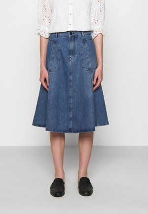 Femme portant une jupe en jean bleue jusqu'aux genoux avec des poches avant, un chemisier blanc en œillet à manches trois-quarts, et des chaussures noires faciles à enfiler.