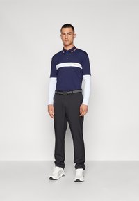 Ναυτικό πόλο μπλουζάκι με άσπρη ρίγα και λογότυπο, συνδυασμένο με μαύρο παντελόνι και άσπρα αθλητικά παπούτσια. Διαθέτει αντίθετο γιακά και μακριά μανίκια.