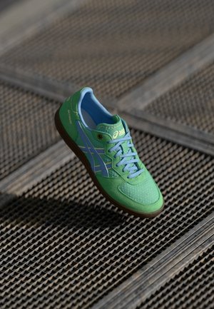 Baskets Asics vertes et bleues avec des lacets bleu clair, coutures visibles et semelle marron, présentées sur un fond de grille métallique.