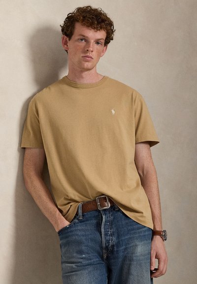 T-shirt in cotone marrone chiaro con collo rotondo e maniche corte. Presenta un piccolo logo bianco sul petto e una vestibilità comoda. Jeans di denim indossati sotto.