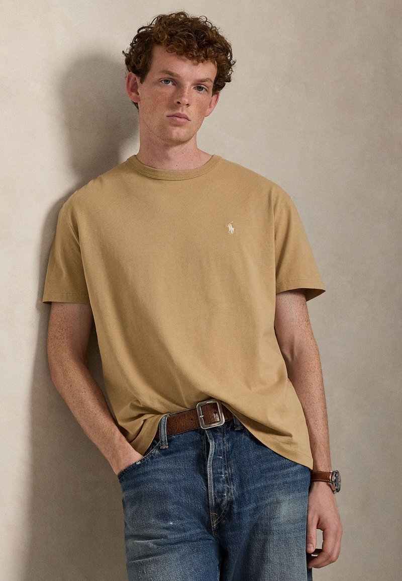 Light brown cotton t-shirt with a round neck and short sleeves. Features a small white logo on the chest and a relaxed fit. Denim jeans worn below.