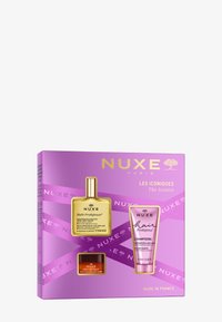 NUXE presentset med en flaska guldolja, en rosa schampotub och en liten röd krämburk. Förpackningen är lila med varumärke och produktinformation.