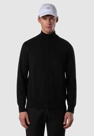 Uomo con berretto bianco e maglione nero con cerniera e tasche laterali, in piedi davanti a uno sfondo grigio chiaro uniforme.