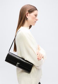 Jeune femme aux cheveux bruns lisses, portant des boucles d'oreilles créoles et un pull blanc, portant un sac à bandoulière noir rectangulaire avec une fermeture argentée.