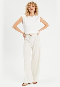 Top blanco sin mangas combinado con pantalones anchos texturados de color beige claro y un cinturón trenzado. Sandalias con acentos decorativos completan el look.