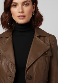 Bruine leren trenchcoat met een uitgesneden kraag, twee knopen en een zachte textuur; gedragen over een zwarte geribde coltrui.