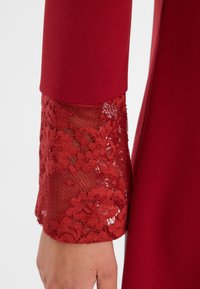Roter Kleidärmel mit einem Oberteil aus festem Stoff und einem kunstvollen unteren Abschnitt aus Spitze mit floralen Mustern, der Netz- und Strukturelemente zeigt.