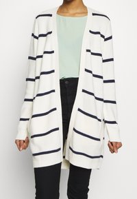 Cárdigan de punto en crema con rayas horizontales en azul marino, con abertura frontal, mangas largas y puños de canalé. Lleva puesto un top verde claro y pantalones negros.