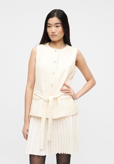 Γυναίκα με μακριά σκούρα μαλλιά που φοράει αμάνικο εκρού γιλέκο με κουμπιά και ζώνη πάνω από πλισέ εκρού φούστα, σε απλό φόντο.